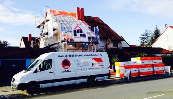 Dachdeckerbetrieb saniert Hausdach mit Gerüst, Lieferwagen und Baumaterialien am Straßenrand.